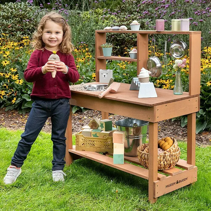 Muddy Buddy Matschküche Mud Starter - 80 x 40 x 90 cm, Holz, Braun - Kind spielt mit der Muddy Buddy Spielküche im Garten, umgeben von Blumen. Diese Spielküche bietet vielfältige Spielmöglichkeiten im Freien.
