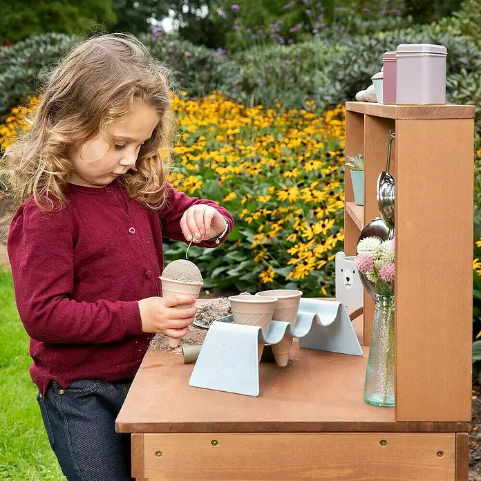 Muddy Buddy Matschküche Mud Starter - 80 x 40 x 90 cm, Holz, Braun - Ein kleines Mädchen spielt an einer hölzernen Spielküche im Garten und füllt Sand in eine Eiswaffel.