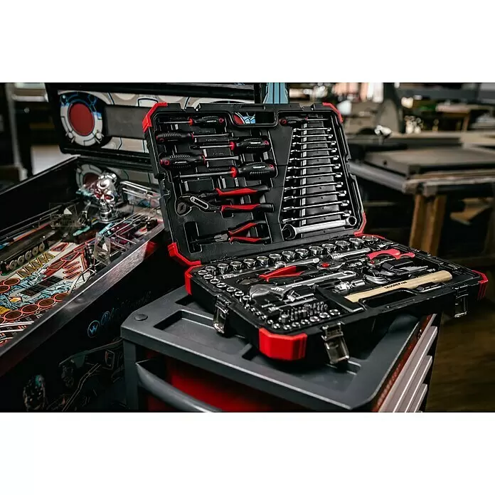 Gedore red Set de llaves de vaso - 100 pzs., Tamaño de accionamiento: ½″ - Un juego de herramientas abierto, negro y rojo, muestra diversas herramientas manuales sobre un carro gris, junto a una máquina de pinball.