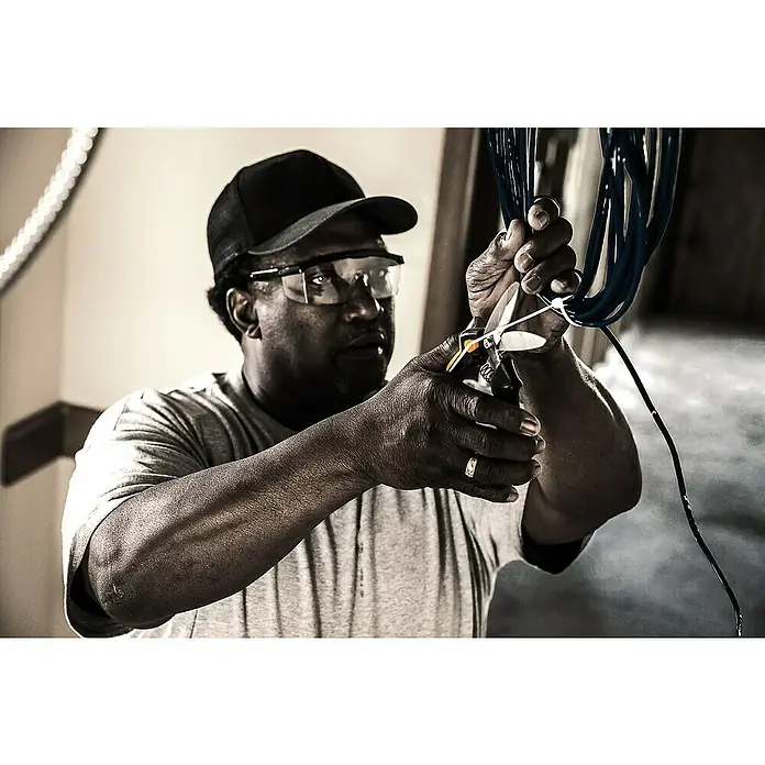 Fiskars PowerArc Tijeras de trabajo 1027204 - Largo: 26 cm, Longitud de corte: 60 mm, Acero inoxidable - Un hombre con gafas de seguridad y gorra corta cables con tijeras, centrándose en la herramienta y el manejo preciso.