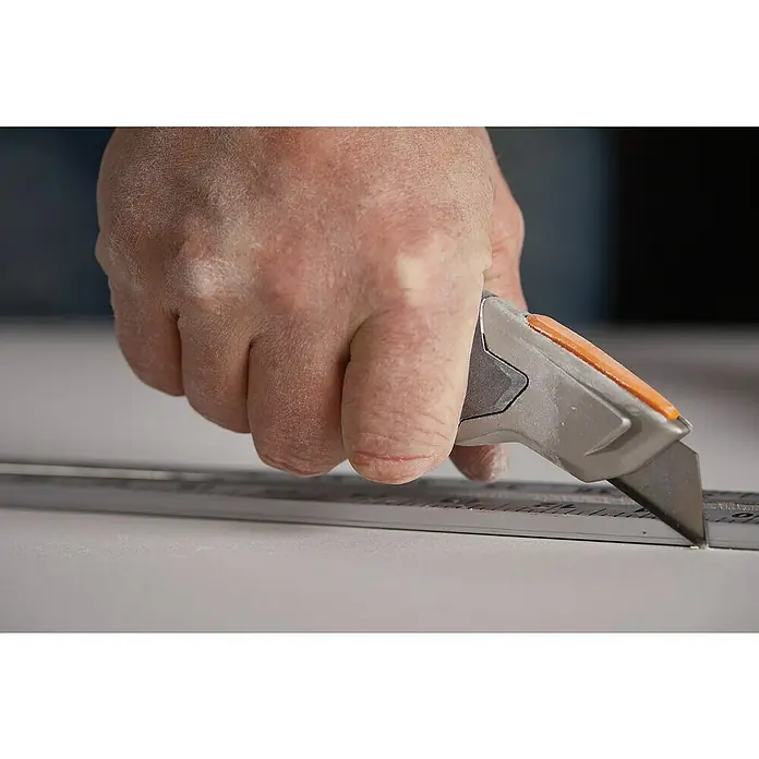 Fiskars CarbonMax Cuttermesser mit feststehender Klinge - 5 Klingenpositionen - Eine Hand schneidet mit einem grauen Cuttermesser und einem orangefarbenen Schieber etwas entlang eines Metalllineals auf einer weißen Oberfläche.