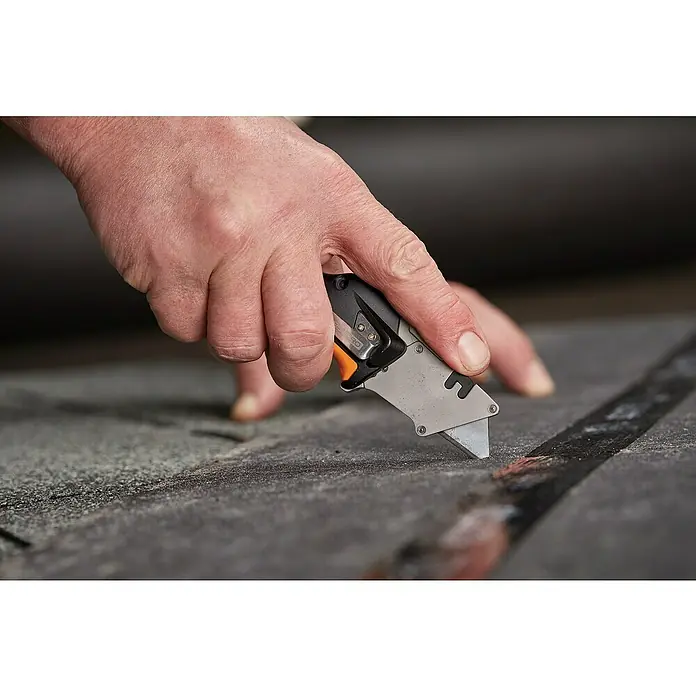 Fiskars CarbonMax Klappmesser - Art Klinge: Trapezklinge, Ergonomischer Griff - Nahaufnahme eines Mannes, der mit einem grauen Cuttermesser mit schwarzem Griff eine dunkle Oberfläche schneidet.