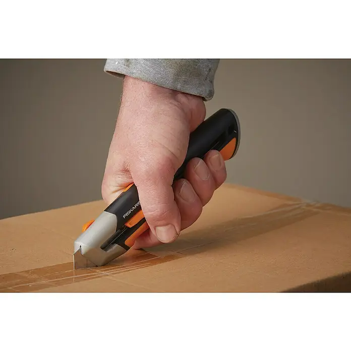Fiskars CarbonMax Cuttermesser - Klingenbreite: 25 mm - Das Bild zeigt einen Kartonschneider von Fiskars, der zum Öffnen eines Kartons verwendet wird. Das Werkzeug liegt sicher in der Hand.