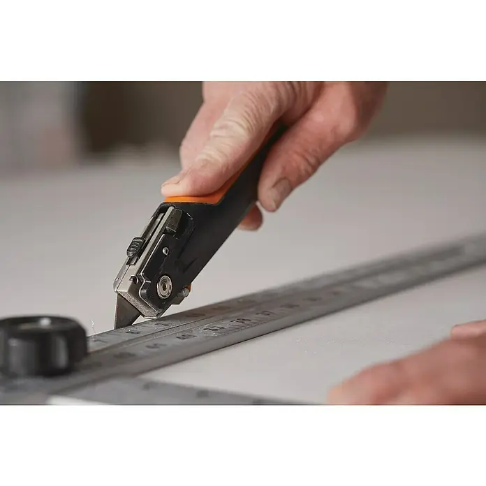 Fiskars CarbonMax Schiebemesser Trockenbau - Art Klinge: Trapezklinge, 3 Klingenpositionen, Schwarz/Orange - Ein Teppichmesser mit orangefarbenem Griff schneidet mit Hilfe eines Metalllineals präzise eine weiße Oberfläche.