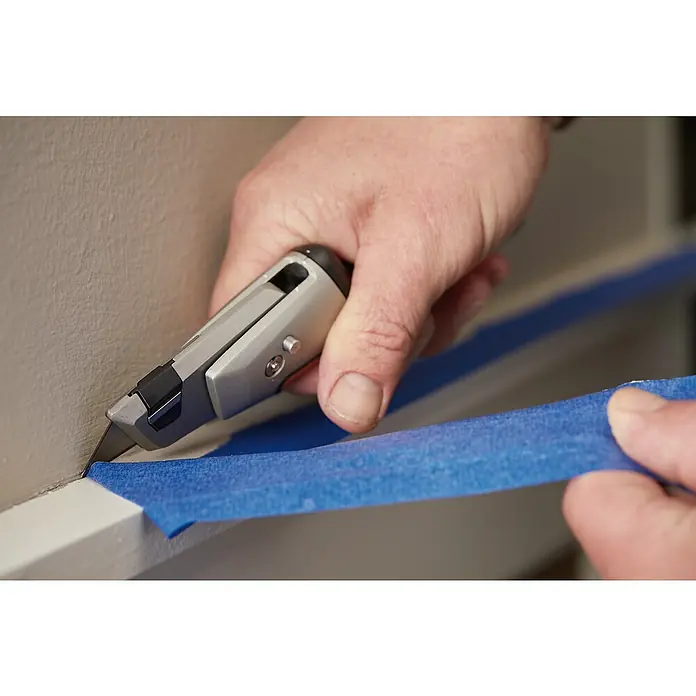 Fiskars CarbonMax Schiebemesser Maler - Art Klinge: Trapezklinge, 3 Klingenpositionen, Schwarz/Orange - Nahaufnahme eines Cuttermessers, das verwendet wird, um Malerkrepp entlang einer weißen Leiste zu schneiden.
