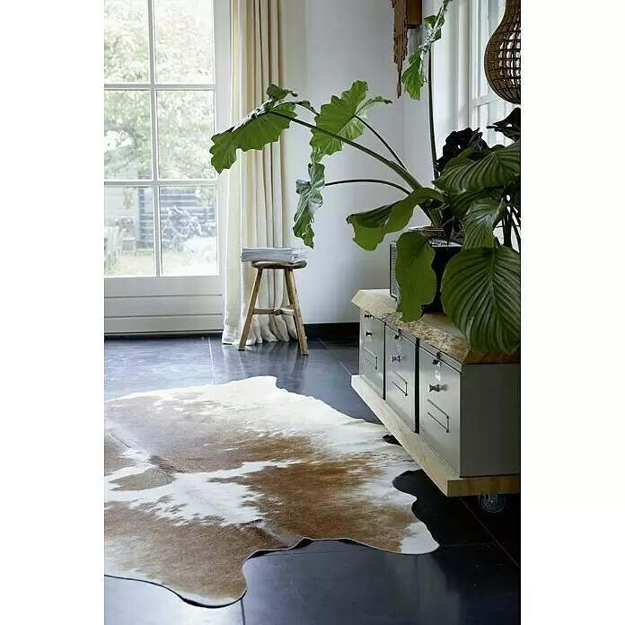 Esbeco Alfombra de piel de vaca - Marrón/Blanco, 100% piel real - Alfombra de piel de vaca marrón y blanca sobre suelo oscuro, parcialmente bajo un mueble, en un espacio acogedor.
