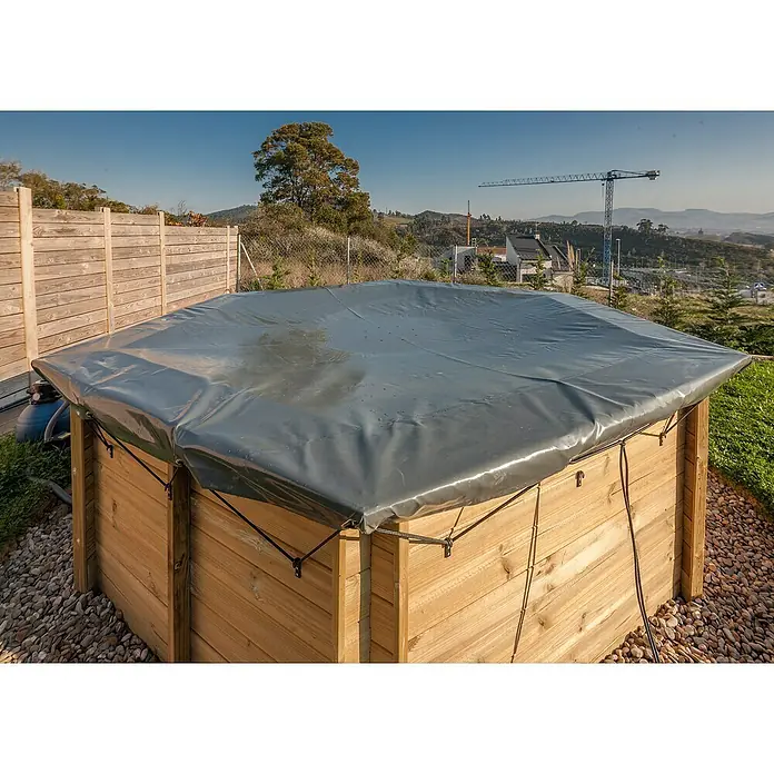 Gre Pool-Abdeckplane Winter Vanille - 580 g/m², Passend für: Pools bis Ø 400 cm - Abgedeckter Holzpool mit grauer Plane, umgeben von Steinen und einem Holzzaun im Hintergrund.