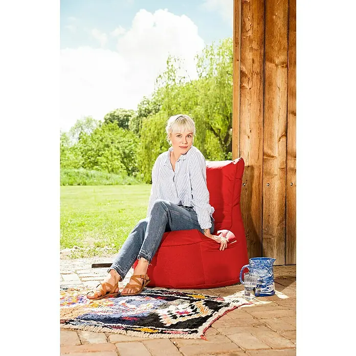 Sitzsack Swing Scuba Set inkl. Dotcom - 95 x 90 cm, Rot, 100 % Polyester/100 % Polyvinylchlorid - Eine Frau sitzt entspannt auf einem roten Sitzsack im Freien, umgeben von Grün und einer rustikalen Holzwand. Der Sitzsack lädt zum Verweilen ein.