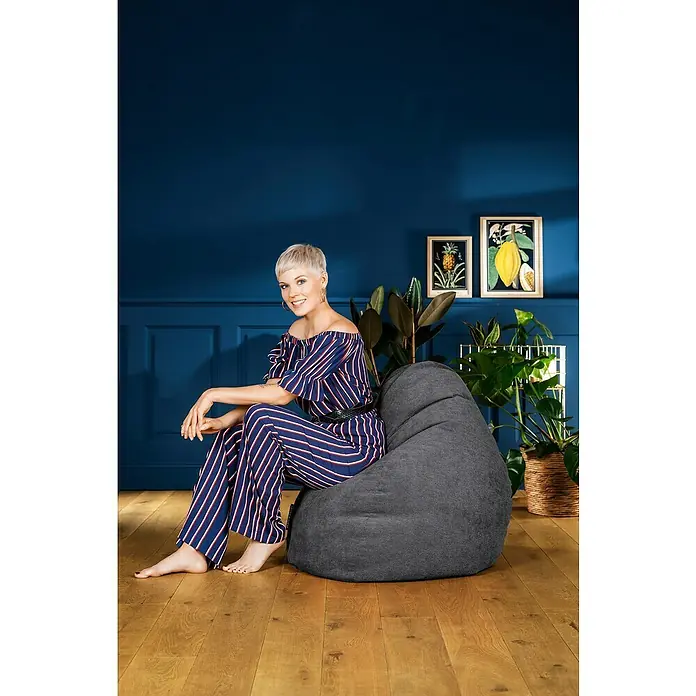 Sitting Point Sitzsack Beanbag Alfa - XL, Anthrazit, 100 % Polyester - Eine Frau sitzt entspannt auf einem grauen Sitzsack vor einer blauen Wand, dekoriert mit Pflanzen und gerahmten Bildern.