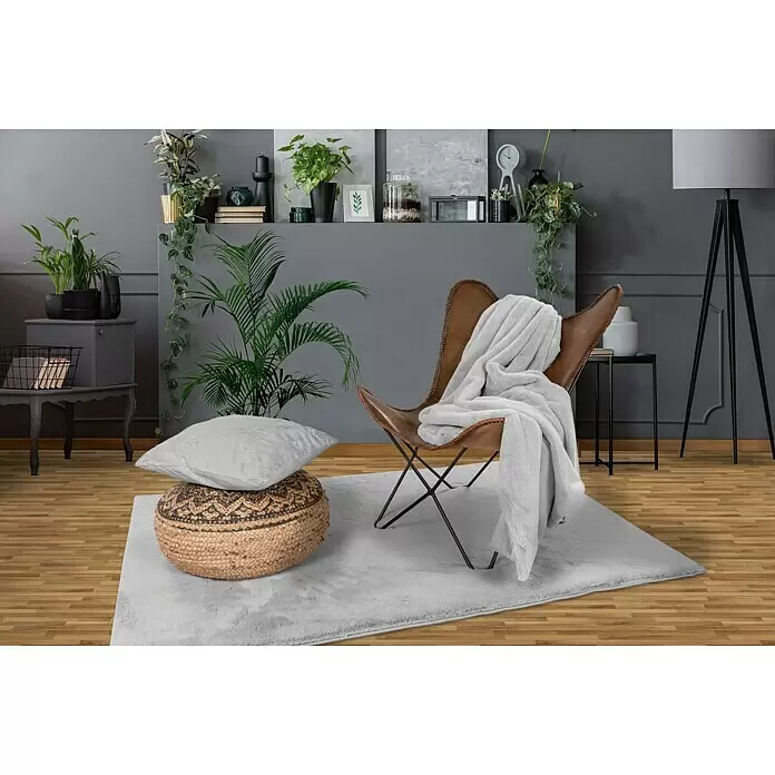 Manta para sofá Happy - 150 x 200 cm, Gris - Una alfombra suave de pelo largo gris claro sobre suelo de madera, en el centro de una sala de estar moderna con sillón de cuero y puf.