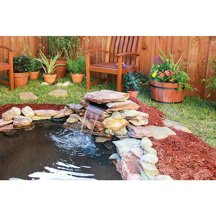 Pontec Einbau-Wasserfall Pondofall - 32,5 x 27 x 16 cm - Ein ruhiger Gartenteich mit einem kleinen Wasserfall, umgeben von Pflanzen in Tontöpfen und einem Holzstuhl, der eine friedliche Gartenszene erzeugt.