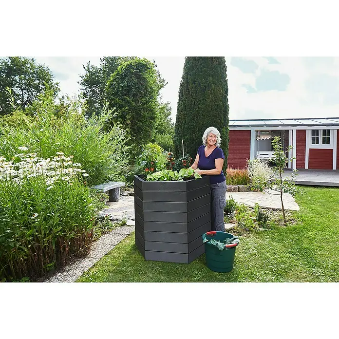 Garantia Hochbeet Ergo Stone - Ø x H: 110 x 25 cm, Kunststoff, Anthrazit - Frau im Garten mit einem sechseckigen, erhöhten Pflanzgefäß aus grauem Holz, in dem Gemüse wächst.