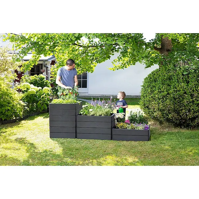 Garantia Hochbeet Ergo Quadro M - 80 x 60 x 25 cm, Kunststoff, Anthrazit, Schieferoptik - Graue Pflanzkästen in gestaffelter Anordnung mit Mann und Kind, die sich um die Bepflanzung kümmern, im Garten vor einem Haus.