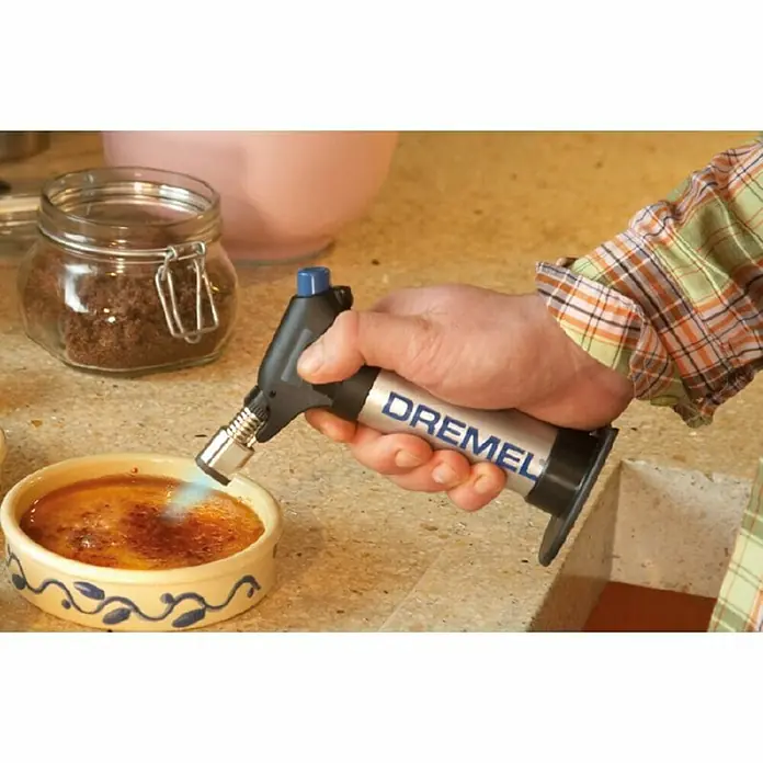 Dremel VersaFlame Unidad de soldadura de alto rendimiento 2200-4 - Apto para: Gas butano - Se está utilizando un soplete Dremel para caramelizar un postre, con un frasco de azúcar moreno y un tazón rosa al fondo.
