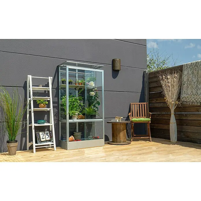 Vitavia Gewächshaus Stad 350 - Außenmaß inkl. Dachüberstand (B x T): 81 x 43 cm, 3 mm, Aluminium, Mit Stahlfundament - Ein gläsernes Gewächshaus voller Pflanzen und Blumen steht auf einer Holzterrasse, daneben eine Leiter mit Dekorationen und eine Sitzgruppe.