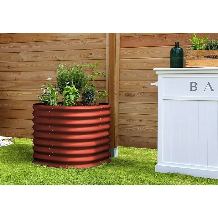 Vitavia Hochbeet Terra 630 - 82 x 82 x 63 cm, Zincalume®, Rubin - Das Bild zeigt einen runden, rotbraunen Pflanzbehälter aus gewelltem Metall mit verschiedenen Pflanzen auf einer grünen Rasenfläche im Garten.