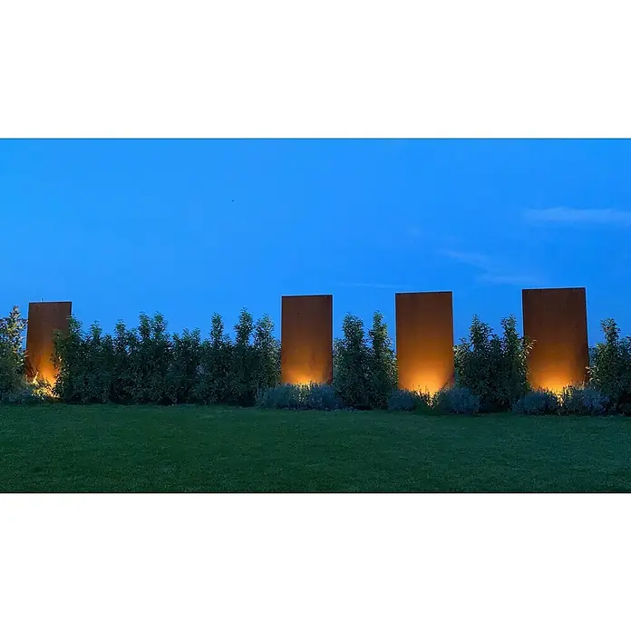 Palatino Space Vertical Sichtschutzelement Uni - 87 x 180 cm, Edelrost, Uni - Gartengestaltung mit vier rostfarbenen Metallpaneelen, die von unten beleuchtet werden, eingebettet in grüne Büsche und Rasen.