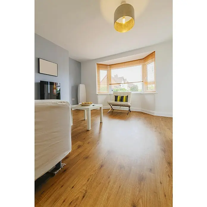 Suelo vinílico SPC Roble Natural - 1.220 x 180 x 5,5 mm, Beige, Resistente al agua - Un salón luminoso con piso de madera, sofá, mesa, sillón junto a la ventana y lámpara de techo. Decoración luminosa y moderna.