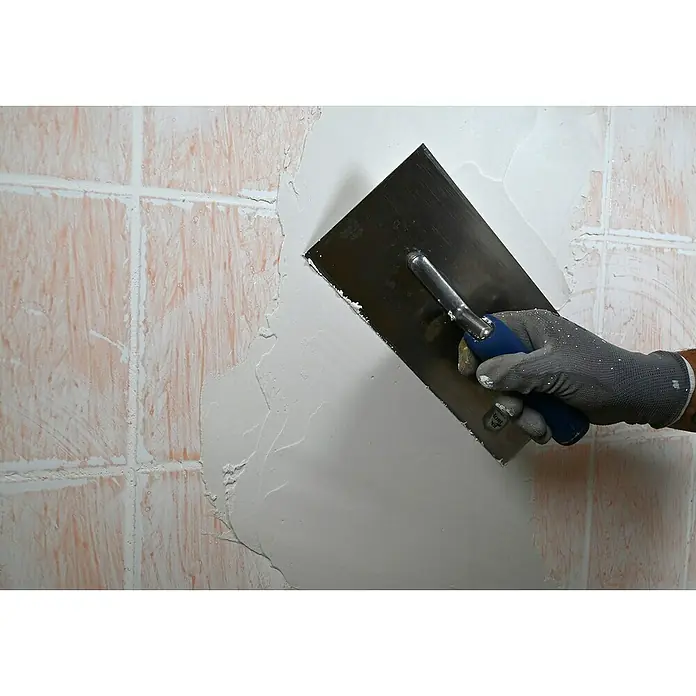Knauf Rotband Flächenspachtel Plus - 4 kg - Eine Hand mit Arbeitshandschuh glättet mit einer Glättkelle Spachtelmasse auf einer Wand mit rosa Fliesen, um eine glatte Oberfläche zu schaffen.