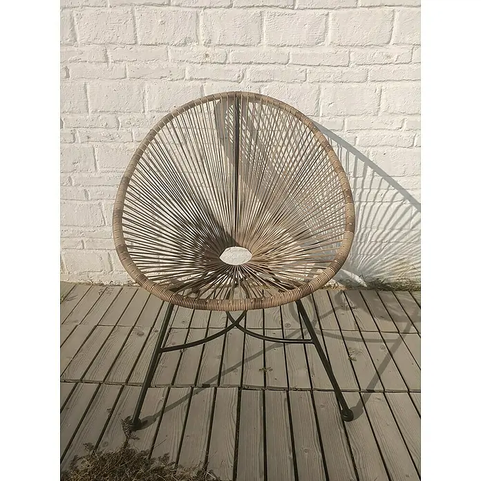 Set de muebles de jardín Fidji - 3 pzs., Acero/Polietileno, Marrón - Una silla Acapulco de mimbre con patas de metal oscuro se encuentra frente a una pared de ladrillo blanco, creando un interesante juego de sombras.