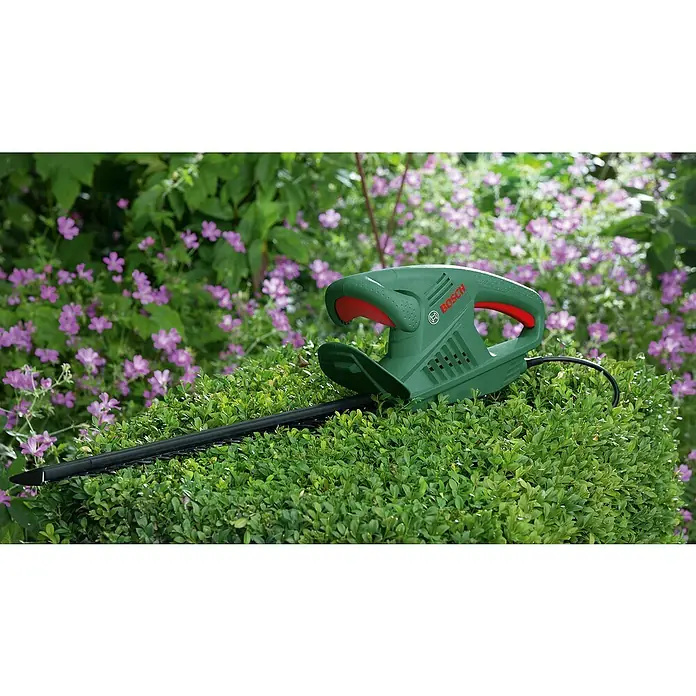 Bosch Cortasetos eléctrico Easyhedgecut 45-16 - 420 W, Longitud de corte: 45 cm - Cortasetos Bosch en uso, rodeado de arbustos verdes y flores moradas. Ideal para jardinería de precisión y mantenimiento de setos.