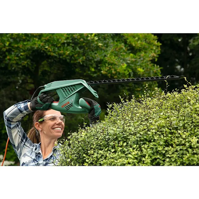 Bosch Elektro-Heckenschere EasyHedgeCut 55 - 450 W, Schnittlänge: 55 cm - Eine Frau benutzt eine Bosch Heckenschere, um eine Hecke zu schneiden, und trägt dabei eine Schutzbrille für die Sicherheit.