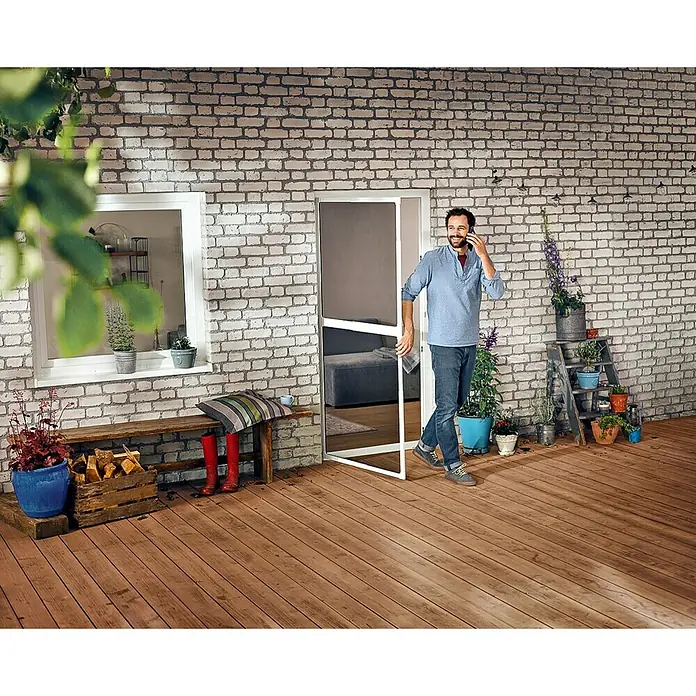 Tesa Insect Stop Puerta con protección contra insectos Falt - Blanco, Montaje de la mosquitera: Pinzas - Un hombre entra a un patio a través de una puerta mosquitera blanca. La puerta impide la entrada de insectos y permite que entre aire fresco.