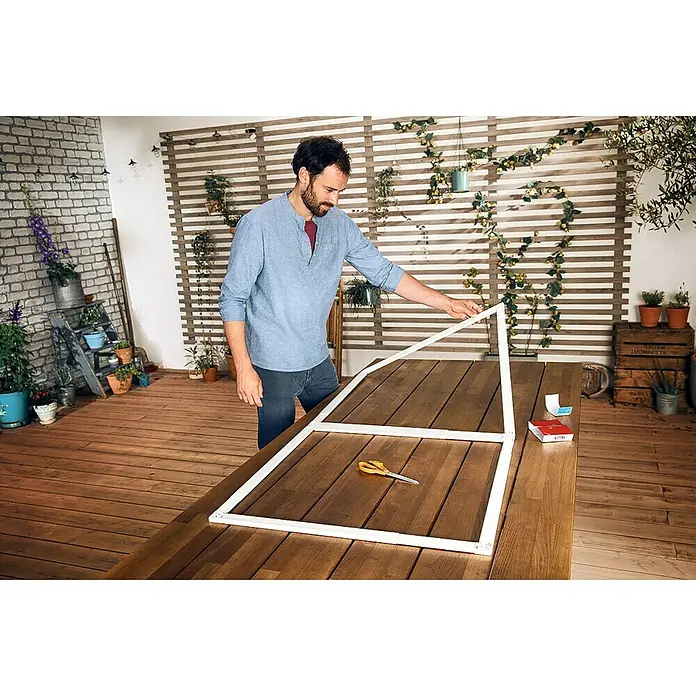 Tesa Insect Stop Puerta con protección contra insectos Falt - Blanco, Montaje de la mosquitera: Pinzas - Un hombre monta un marco blanco sobre una mesa de madera al aire libre, rodeado de plantas. Unas tijeras y una pequeña herramienta están sobre la mesa.