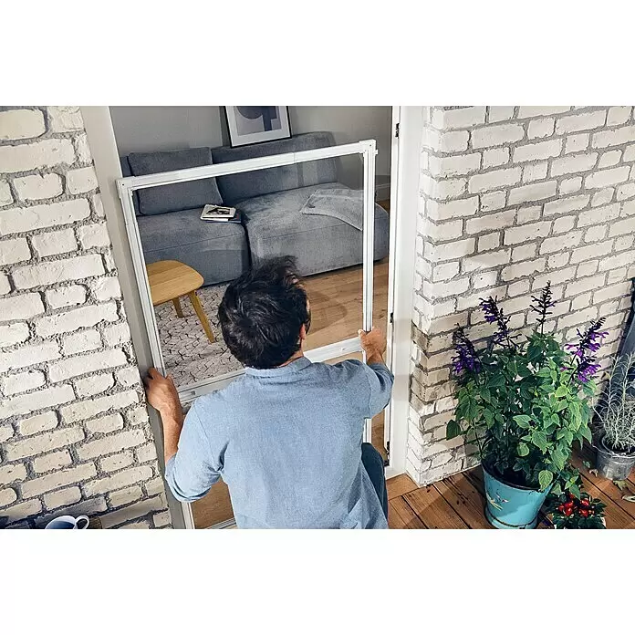 Tesa Insect Stop Puerta con protección contra insectos Falt - Blanco, Montaje de la mosquitera: Pinzas - Hombre instalando un marco blanco de puerta mosquitera en una entrada, mostrando su uso como barrera entre interior y patio.