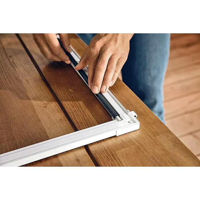 Tesa Insect Stop Puerta con protección contra insectos Falt - Blanco, Montaje de la mosquitera: Pinzas - Primer plano de una persona montando un marco de mosquitera blanco sobre una mesa de madera, fijando el sello al marco.