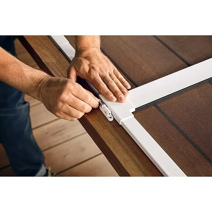 Tesa Insect Stop Puerta con protección contra insectos Falt - Blanco, Montaje de la mosquitera: Pinzas - Un hombre instala una mosquitera con marco blanco sobre una mesa de madera. La pantalla es marrón y de malla fina.