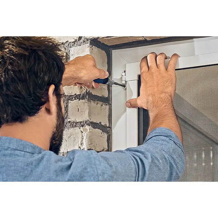 Tesa Insect Stop Puerta con protección contra insectos Falt - Blanco, Montaje de la mosquitera: Pinzas - Un hombre repara una ventana con un destornillador. Asegura las bisagras al marco blanco, garantizando la funcionalidad.