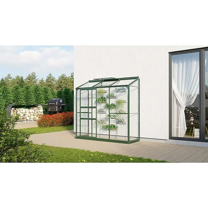Vitavia Gewächshaus Styx 1300 - Außenmaß inkl. Dachüberstand (B x T): 192,8 x 69 cm, 3 mm, Smaragd - Ein Gewächshaus aus Glas mit grünen Rahmen, angelehnt an eine weiße Hauswand, voller Pflanzen in einem Garten mit Grill.
