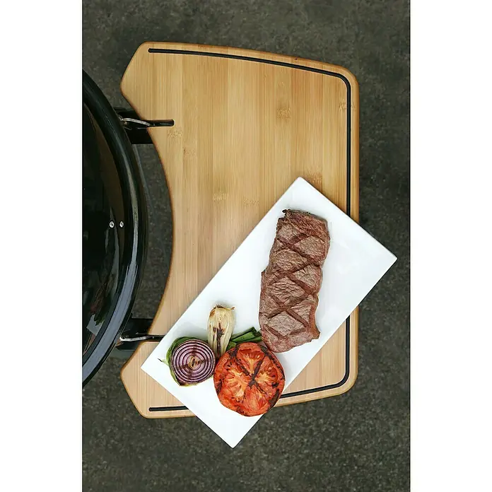 Outdoorchef Seitentisch Bambus 570 - Holz, Passend für: Outdoorchef Kugelgrills Ø 57 cm - Nahaufnahme eines auf einem Schneidebrett angerichteten Grillgerichts mit Steak, Tomaten und Zwiebeln auf einem weißen Teller.