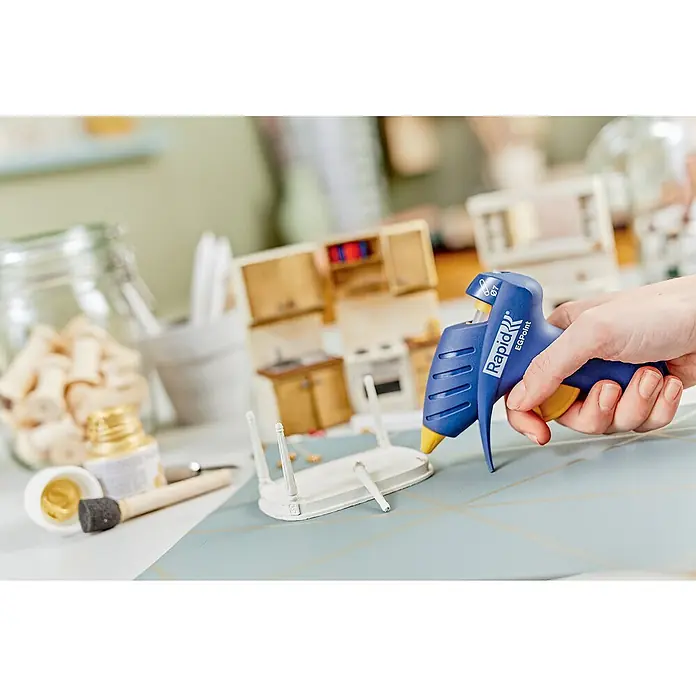 Rapid Pistola termoencoladora EG Point - Diámetro cartucho de cola: 7 mm, 80 W - Se utiliza una pistola de pegamento azul Rapid EGPoint para ensamblar un mueble en miniatura con materiales de manualidades al fondo.