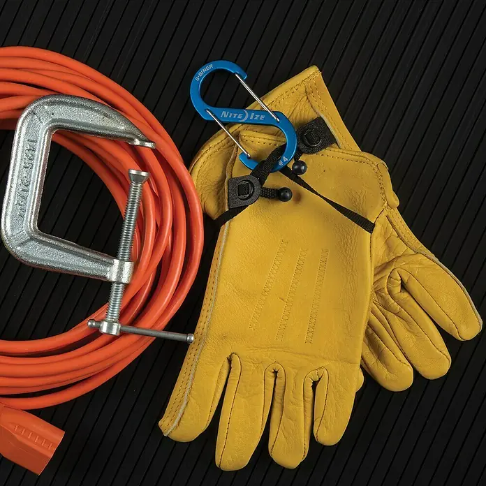 Nite Ize Mosquetón doble S-Biner - 3 ud., Metal - Primer plano de una abrazadera para tubos, cable naranja, guantes de trabajo y un mosquetón azul Nite Ize S-Biner sobre una superficie negra.