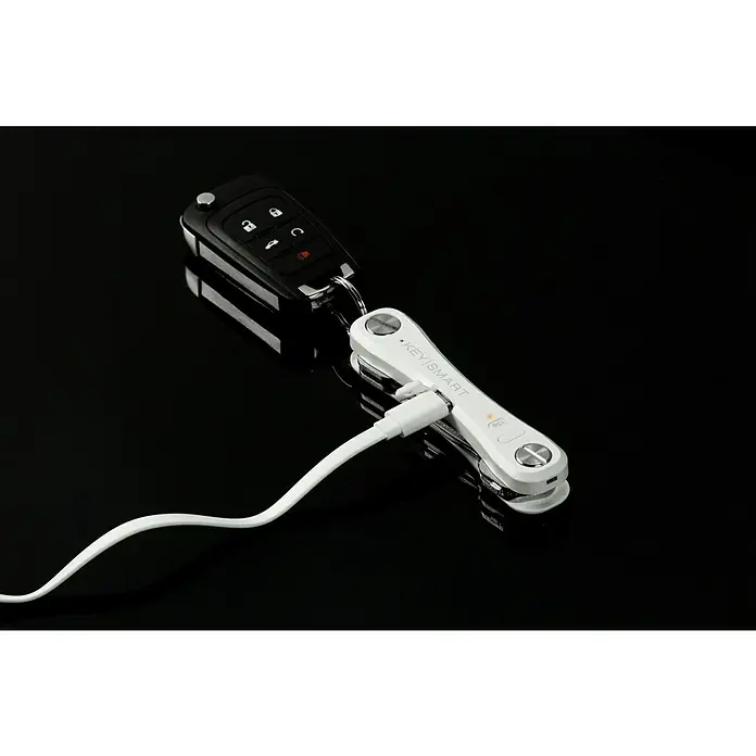 Nite Ize Llavero Key Smart Pro - Largo: 9,2 cm, Acero, 14 Llaves - Primer plano de un buscador de llaves KeySmart Pro con una llave de coche y un cable de carga sobre un fondo negro brillante.