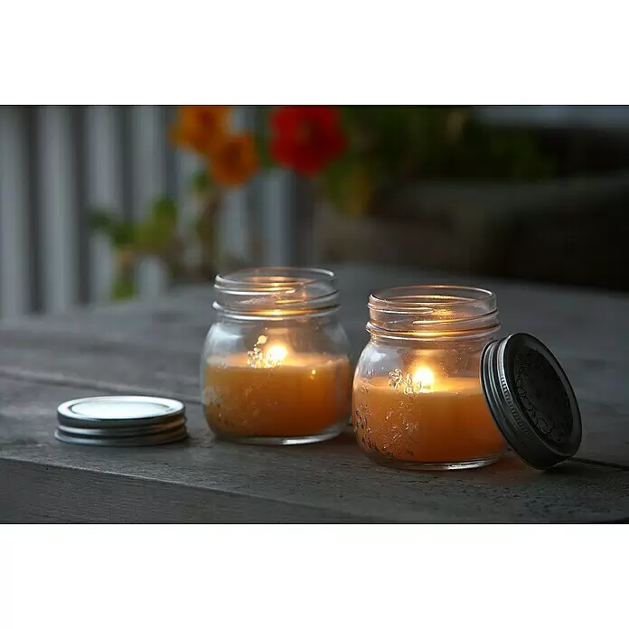 Duftkerze Citronella Potty im Glas - Ø x H: 8 x 9 cm, Citronella - Zwei brennende Kerzen in Gläsern mit orangefarbenem Wachs und Metalldeckeln auf einem Holztisch. Ein Deckel liegt daneben, der andere lehnt.
