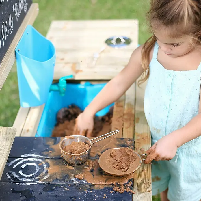 TP Toys Matschküche Deluxe - 40 x 136 x 96 cm, Holz, Natur - Mädchen spielt mit einer Matschküche, umgeben von rustikalem Holz und blauem Zubehör, ideal für kreative Outdoor-Aktivitäten.