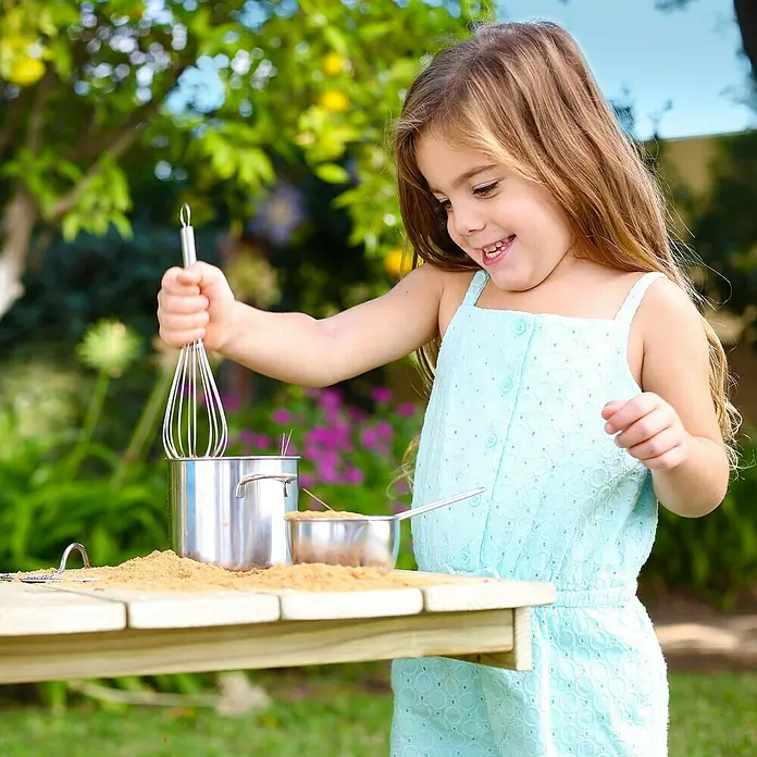 TP Toys Matschküche Deluxe - 40 x 136 x 96 cm, Holz, Natur - Ein kleines Mädchen spielt mit Spielzeugtöpfen und einem Schneebesen im Garten und ahmt das Kochen nach.