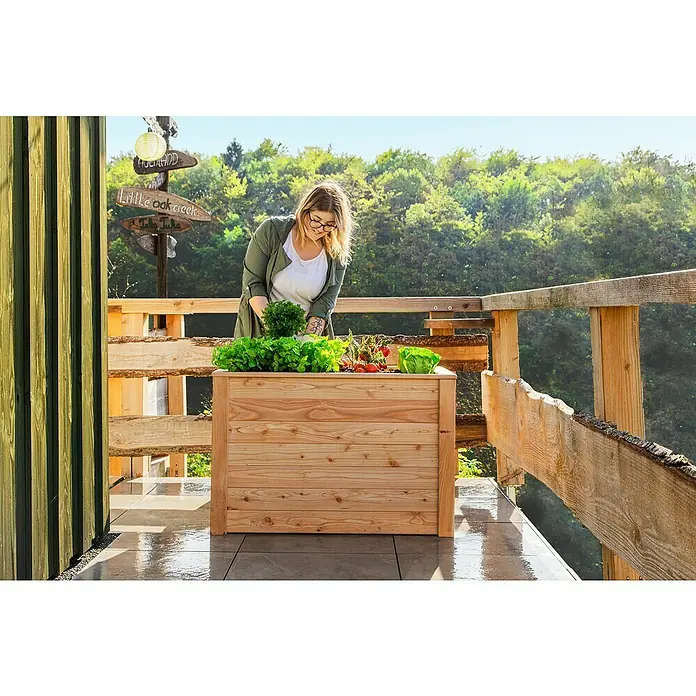 Westmann Hochbeet Lärche 100 - 100 x 65 x 66 cm, Holz, Natur - Eine Frau bepflanzt einen erhöhten Holzpflanzkasten mit Salat und Tomaten auf einer sonnigen Terrasse mit Holzbalkongeländer.