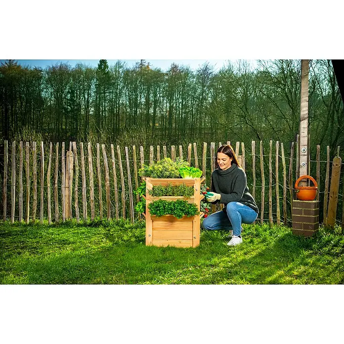 Westmann Hochbeet Premium L - 65 x 65 x 73 cm, Holz, Natur - Eine Frau pflegt einen Holz-Pflanzturm mit drei Ebenen voller frischer Kräuter und Salate in einem Garten.