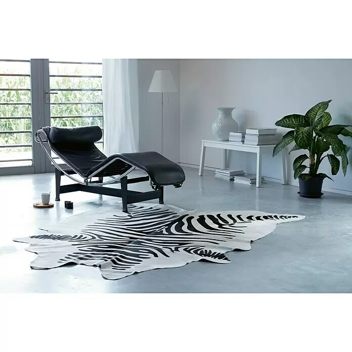 Esbeco Alfombra de piel de vaca - Marrón/Blanco, 100% piel real - Una alfombra de piel de vaca con estampado de cebra en blanco y negro sobre suelo gris claro, parcialmente bajo una chaise longue negra en una habitación moderna.