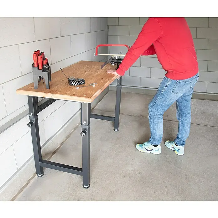 Rikta Werkbank Höhenverstellbar - L x B: 60 x 135 cm, Arbeitshöhe: 105 cm, Belastbarkeit: 200 kg - Eine höhenverstellbare Werkbank mit Werkzeugen und einer Person, die sägt. Ideal für Heimwerkerarbeiten.