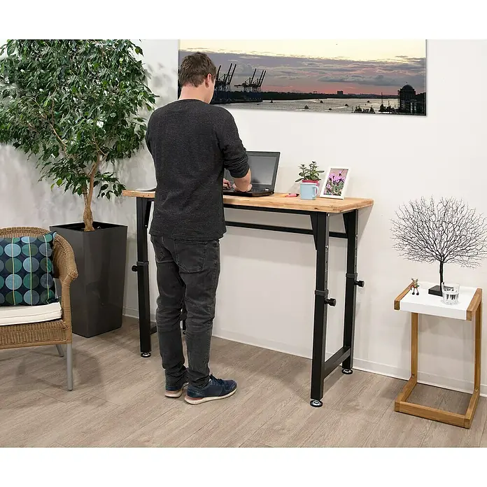 Rikta Werkbank Höhenverstellbar - L x B: 60 x 135 cm, Arbeitshöhe: 105 cm, Belastbarkeit: 200 kg - Ein höhenverstellbarer Schreibtisch aus Holz mit schwarzem Metallgestell steht in einem hellen Raum mit einer Person, die daran arbeitet.