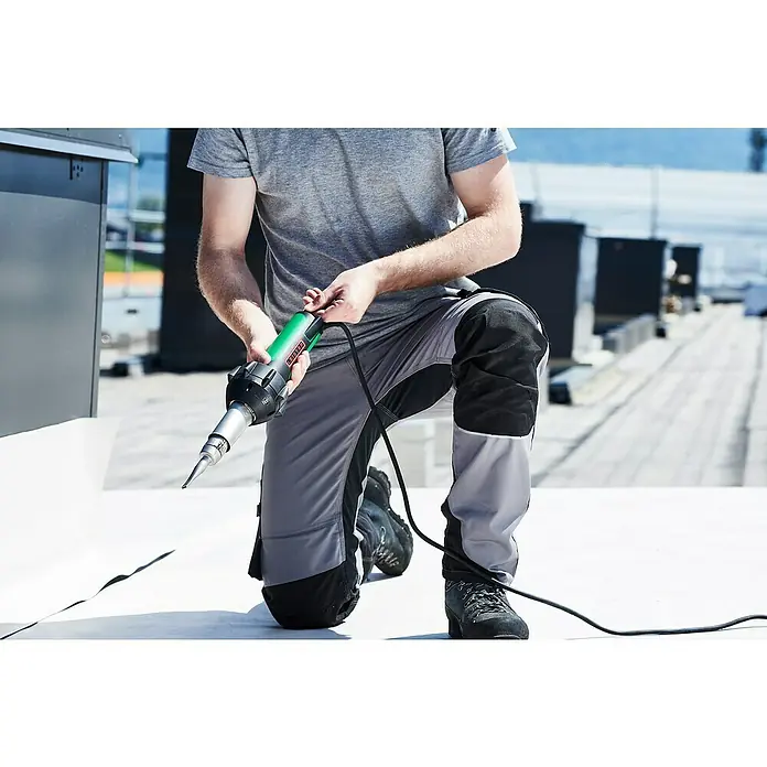 Leister Heißluftgebläse Triac ST - Luftstromregulierung: Keine, Temperatureinstellung: Stufenlos, Analog - Ein Handwerker kniet auf einem Dach und arbeitet mit einem LEISTER Heißluftschweißgerät, um Materialien fachmännisch zu verbinden.