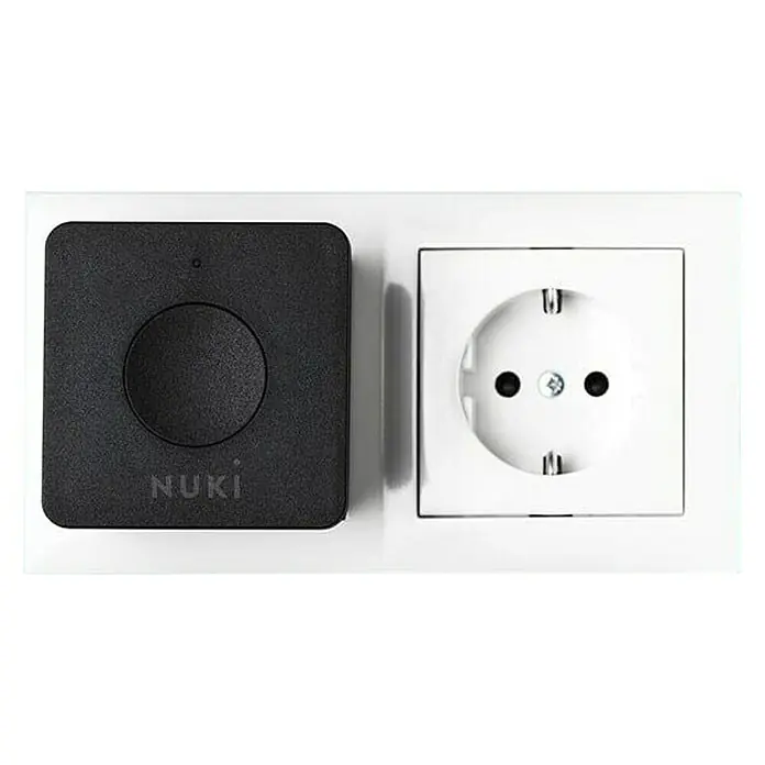 Nuki Módulo transmisor inalámbrico Bridge - Específico para: Cerradura electrónica Nuki, Negro - Primer plano de un dispositivo Nuki Smart Lock junto a una toma de corriente, ambos montados en una pared blanca. El dispositivo Nuki es cuadrado y negro.