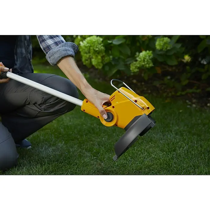 Stiga 100er Serie Akku-Rasentrimmer GT 100e - Set - 20 V, 2 Ah, Li-Ionen, 1 Akku, Schnittbreite Faden: 25 cm - Eine Person hält einen gelben Rasentrimmer mit einem grauen Schaft über grünem Gras. Der Trimmer ist bereit für den Einsatz im Garten.