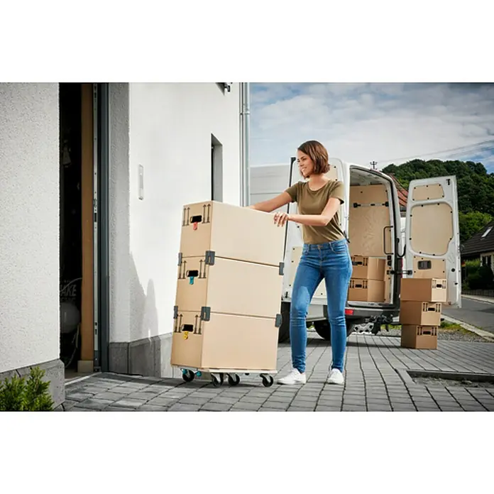 Wolfcraft Base con ruedas FT300 - L x An x Al: 565 x 390 x 120 mm, Capacidad de carga: 200 kg, Madera - Una mujer está empujando una pila de cajas de mudanza en un carrito hacia un garaje con una furgoneta aparcada al fondo.