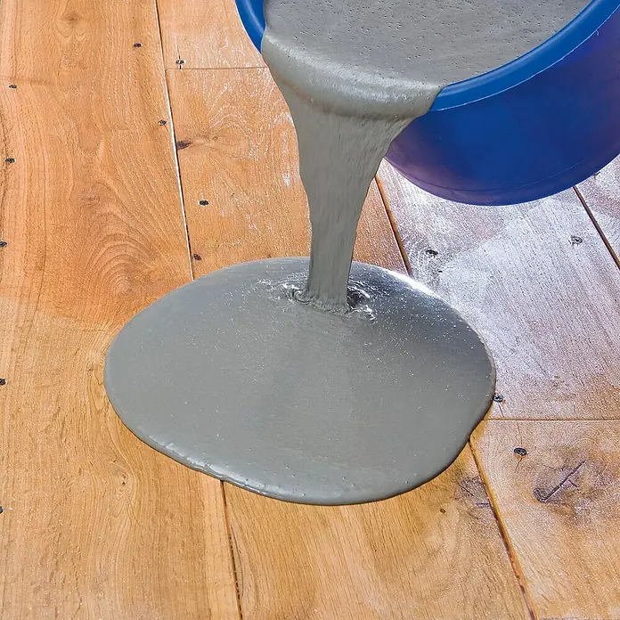 Knauf Fließspachtel Faserflex - 5 kg, Schichtdicke: 2 mm - 15 mm - Der Ausgleichsmörtel wird aus einem blauen Eimer auf einen Holzboden gegossen. Perfekt für die Bodenvorbereitung und Renovierungsarbeiten.